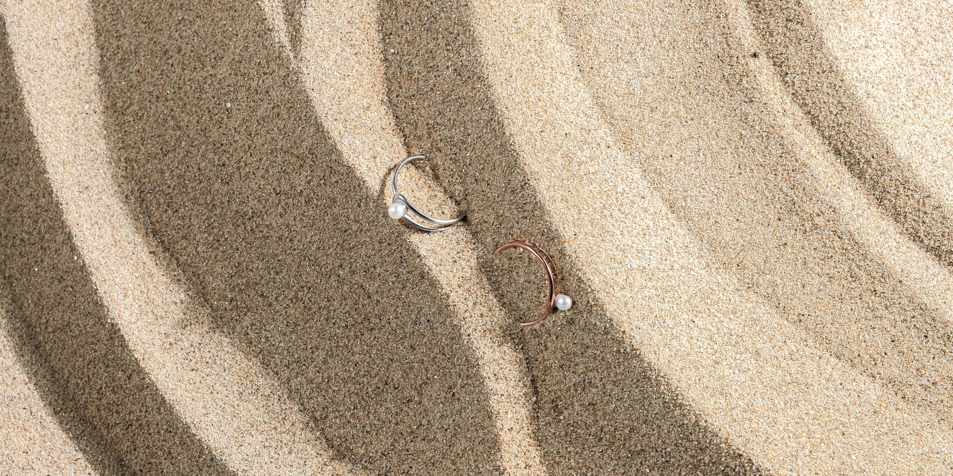 Die Cachet Ringe in Silber und Roségold liegen auf feinem Sand, vollständige Draufsicht.
